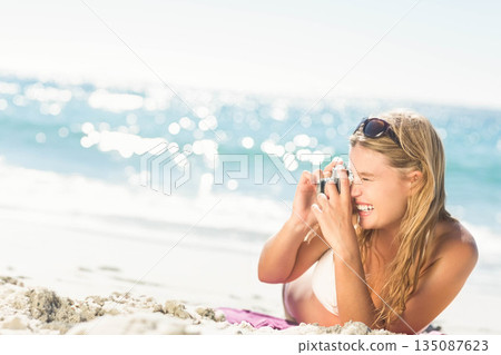 Woman lying on white sand at shoreline wearing bikini top and holding compact camera, copy space 135087623
