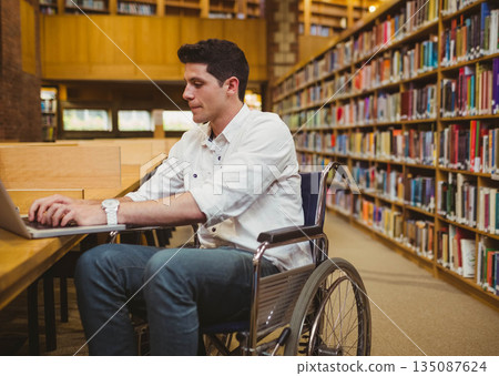 Man in wheelchair typing on laptop at wooden study table inside library with bookshelves Man in wheelchair typing on laptop at wooden study table inside library with bookshelves 135087624