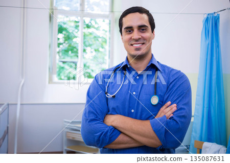 Middle-aged man standing with arms crossed in medical exam room wearing stethoscope by window 135087631