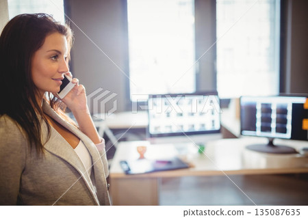 Woman wearing blazer standing by office desk with monitors holding smartphone to ear, copy space 135087635