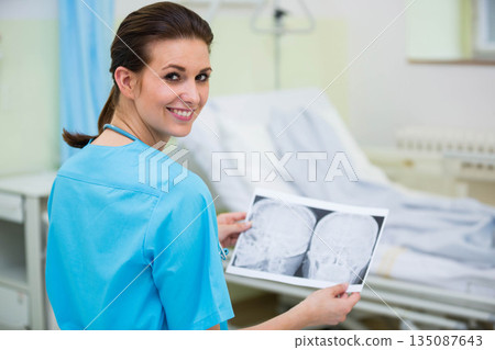 Female nurse wearing blue scrubs holding and examining skull xray films beside bed in care room 135087643