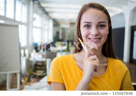 Woman standing in open-plan office holding pen at chin and smiling near flip chart, copy space 135087646