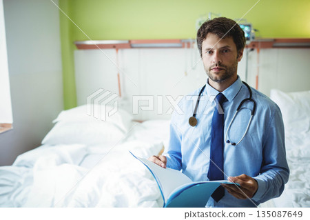 Male doctor standing beside hospital bed reviewing patient file using stethoscope, copy space Male doctor standing beside hospital bed reviewing patient file using stethoscope, copy space 135087649