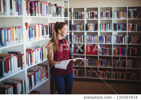 Teenage girl standing in library aisle holding open book and reading among bookshelves, copy space Teenage girl standing in library aisle holding open book and reading among bookshelves, copy space 135087658