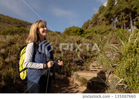 Female hiker standing on trail using trekking poles with yellow backpack and camera, copy space 135087660