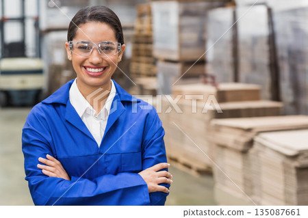African American woman wearing coverall and goggles standing in warehouse with pallets and forklift 135087661