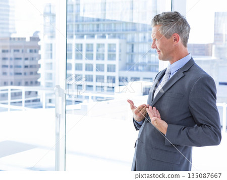 Man in suit standing by window overlooking balcony railing gesturing at city skyline, copy space 135087667