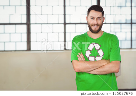 Man in 30s standing with arms crossed by large window wearing green recycling shirt, copy space 135087676