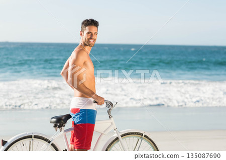 Shirtless man wearing red white blue trunks smiling on beach next to cruiser bicycle, glancing back Shirtless man wearing red white blue trunks smiling on beach next to cruiser bicycle, glancing back 135087690