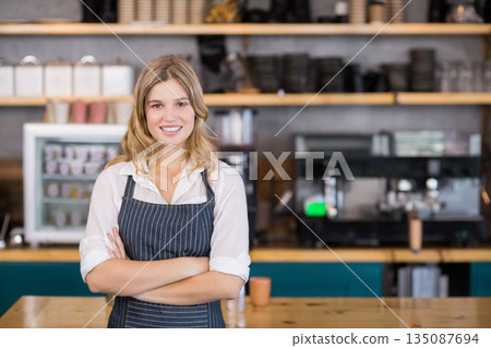Female barista standing behind coffee bar wearing apron and posing by espresso machine in cafe 135087694