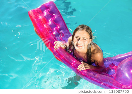 Woman in swimsuit leaning on translucent pink pool lounger and smiling in outdoor pool Woman in swimsuit leaning on translucent pink pool lounger and smiling in outdoor pool 135087700