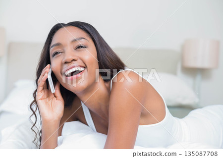 African American woman talking on smartphone while lying on bed in bedroom with pillows and lamp 135087708