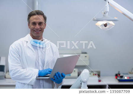 Male dentist wearing white lab coat holding laptop, smiling in dental clinic with lamp, copy space Male dentist wearing white lab coat holding laptop, smiling in dental clinic with lamp, copy space 135087709