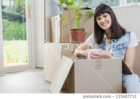 Woman wearing vest sitting on floor in room unpacking moving boxes with bubble wrap, copy space 135087711