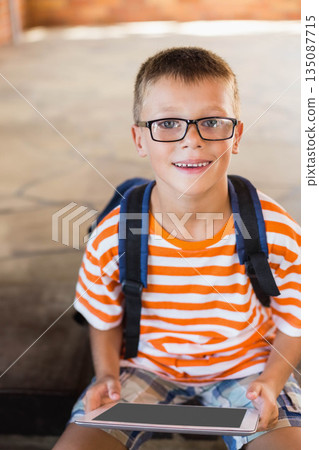 School-aged boy with eyeglasses sitting on tiled step in covered patio holding tablet with backpack School-aged boy with eyeglasses sitting on tiled step in covered patio holding tablet with backpack 135087715