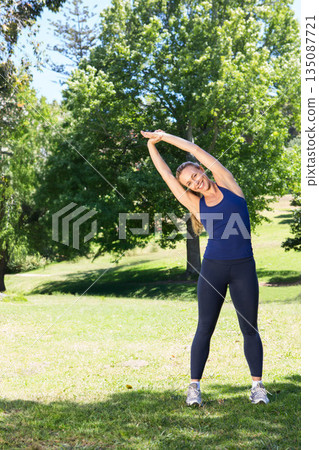 Woman performing side bend stretch on park lawn wearing navy tank top black leggings grey shoes 135087721