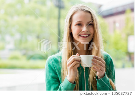 Smiling teenage girl wearing green zip-up sweatshirt standing on cafe patio holding white mug 135087727