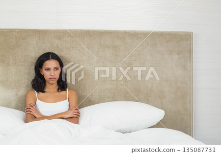 African American woman sitting on bed in bedroom, crossing arms before beige headboard and pillows African American woman sitting on bed in bedroom, crossing arms before beige headboard and pillows 135087731