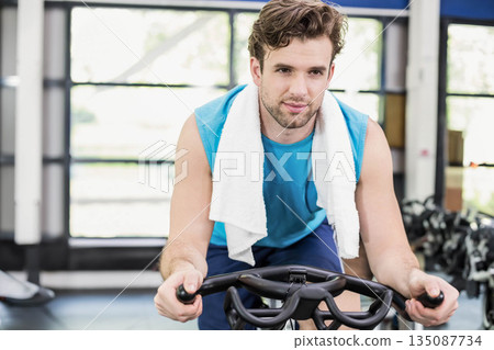 Adult male pedaling stationary bike near weight racks in gym wearing sleeveless teal top and towel Adult male pedaling stationary bike near weight racks in gym wearing sleeveless teal top and towel 135087734