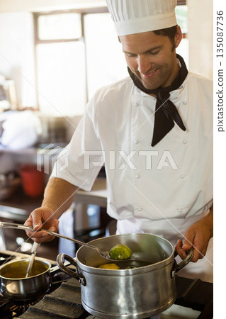Male chef wearing uniform stirring green vegetables in steel pot on gas stove in restaurant kitchen 135087736