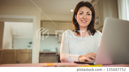 Woman typing on laptop at bright kitchen workspace with light wood cabinets behind, copy space Woman typing on laptop at bright kitchen workspace with light wood cabinets behind, copy space 135087752