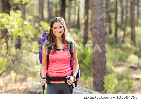 Woman hiker trekking on pine woodland trail wearing athletic gear and carrying backpack and poles 135087753