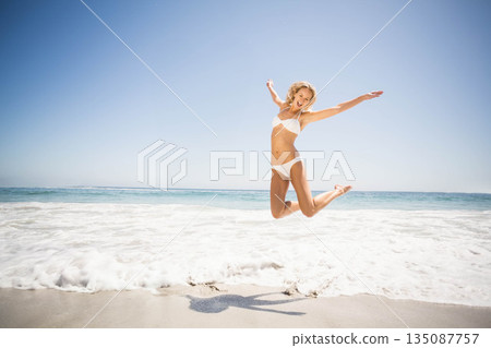 Female wearing white bikini jumping on sandy shoreline with foamy waves under clear sky Female wearing white bikini jumping on sandy shoreline with foamy waves under clear sky 135087757