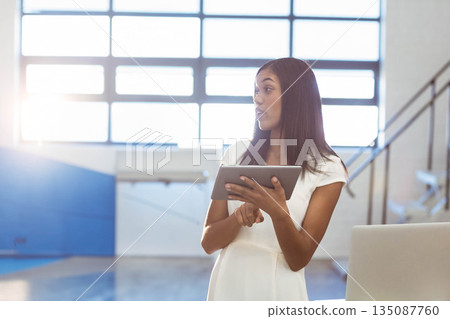 Woman presenting data on tablet in white dress while standing in gymnasium with laptop and railing 135087760