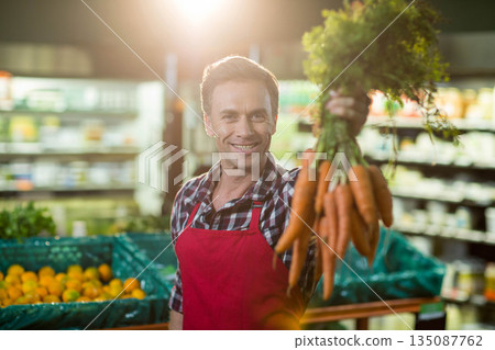 Male grocery worker wearing red apron holding bunch of carrots toward camera in produce aisle Male grocery worker wearing red apron holding bunch of carrots toward camera in produce aisle 135087762