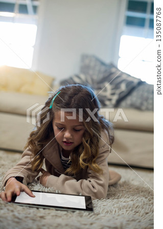 Girl child lying on plush carpet near sofa in living room using tablet with hair clip Girl child lying on plush carpet near sofa in living room using tablet with hair clip 135087768
