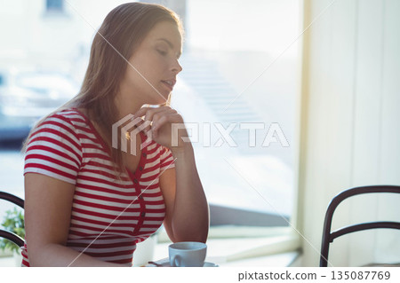 Woman sitting at cafe table by window resting chin on hand gazing over white coffee cup Woman sitting at cafe table by window resting chin on hand gazing over white coffee cup 135087769
