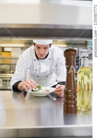 Male chef in chef jacket leaning over kitchen counter, garnishing salad with herbs and pepper mill Male chef in chef jacket leaning over kitchen counter, garnishing salad with herbs and pepper mill 135087771