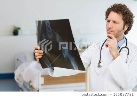 Male doctor wearing lab coat, stethoscope, holding X-ray in exam room with hospital bed, copy space Male doctor wearing lab coat, stethoscope, holding X-ray in exam room with hospital bed, copy space 135087781