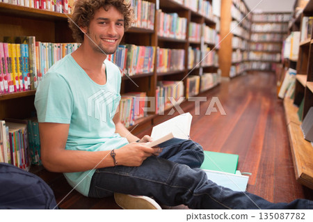 Male student reading open book sitting on floor with backpack beside tall bookshelves, copy space Male student reading open book sitting on floor with backpack beside tall bookshelves, copy space 135087782