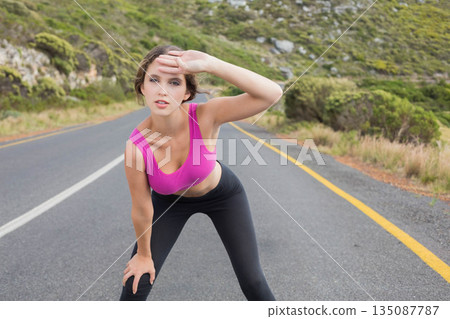 Female runner wearing fuchsia sportswear pausing on mountain highway beside white, yellow lines Female runner wearing fuchsia sportswear pausing on mountain highway beside white, yellow lines 135087787