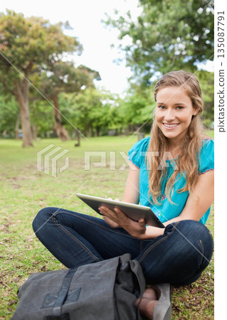 Woman sitting cross-legged on grass in park holding tablet computer beside backpack, copy space 135087791