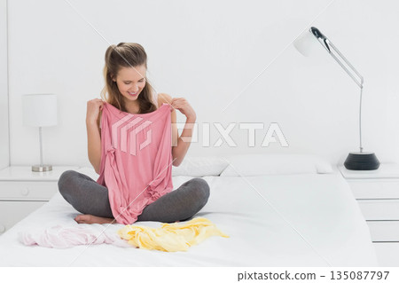 Woman holding pink top while comparing pale pink and yellow shirts on bed in minimalist bedroom Woman holding pink top while comparing pale pink and yellow shirts on bed in minimalist bedroom 135087797