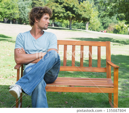 Man sitting on wooden bench on grassy park lawn folding arms and gazing left, copy space 135087804