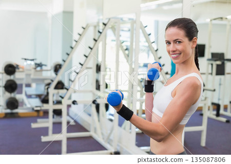 Woman lifting blue dumbbells wearing white sports bra at gym weight rack and mirrors, copy space Woman lifting blue dumbbells wearing white sports bra at gym weight rack and mirrors, copy space 135087806