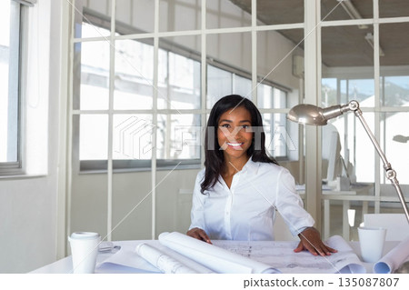African American woman reviewing architectural blueprints at office desk with coffee cup and lamp African American woman reviewing architectural blueprints at office desk with coffee cup and lamp 135087807