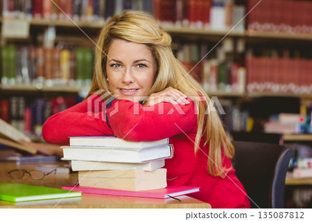 Woman wearing red sweater leaning on textbooks at table in library with notebook and eyeglasses 135087812