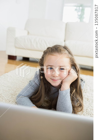 Female child lying on stomach propping head on off-white rug in living room viewing laptop Female child lying on stomach propping head on off-white rug in living room viewing laptop 135087817