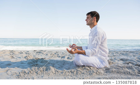 Man sitting cross-legged meditating at sandy shoreline facing ocean under clear sky, copy space 135087818
