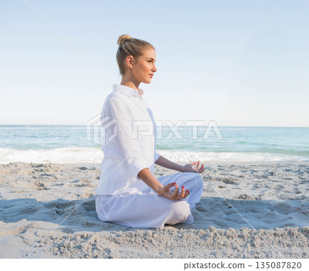 Woman meditating cross-legged on sandy beach by ocean wearing white linen outfit 135087820