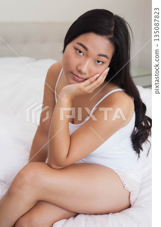 Korean woman sitting cross-legged on bed in pink camisole with white linens and glass nightstand 135087823