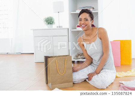 African American woman smiling while handling brown paper bag sitting in bedroom with gift bags 135087824