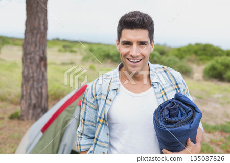 Male in his twenties standing at campsite holding rolled-up blue sleeping bag beside green red tent 135087826