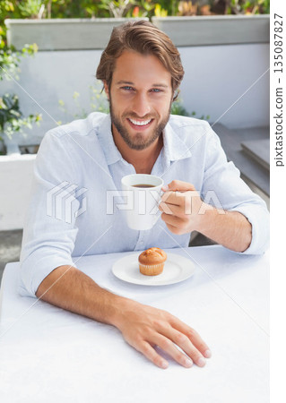 Man smiling while sitting at outdoor cafe terrace on white tablecloth holding coffee mug and muffin 135087827