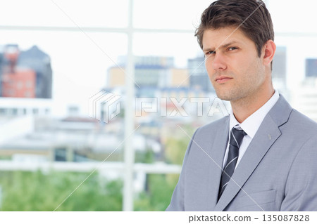 Thirty-something male standing by grid window at office wearing gray suit, striped tie, copy space 135087828