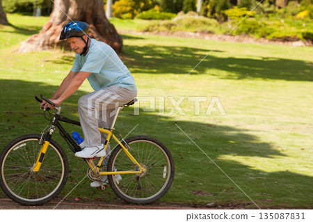 Senior man riding yellow bicycle with blue helmet, water bottle on frame in park, copy space 135087831